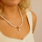 A person wearing the WJW Semi Precious Natural Stone Charm Keeper Necklace, which features a white beaded strand with an 18K Gold PVD coated ring and bead pendant, dressed in a cream-colored tank top with scalloped trim.