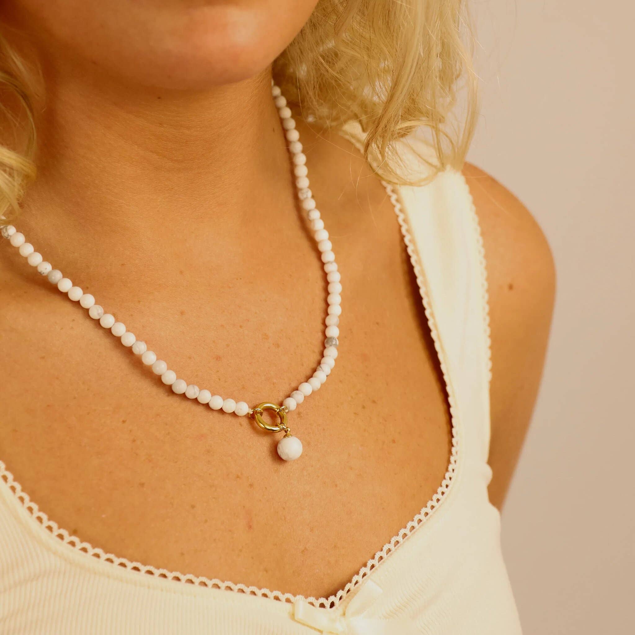 A person wearing the WJW Semi Precious Natural Stone Charm Keeper Necklace, which features a white beaded strand with an 18K Gold PVD coated ring and bead pendant, dressed in a cream-colored tank top with scalloped trim.