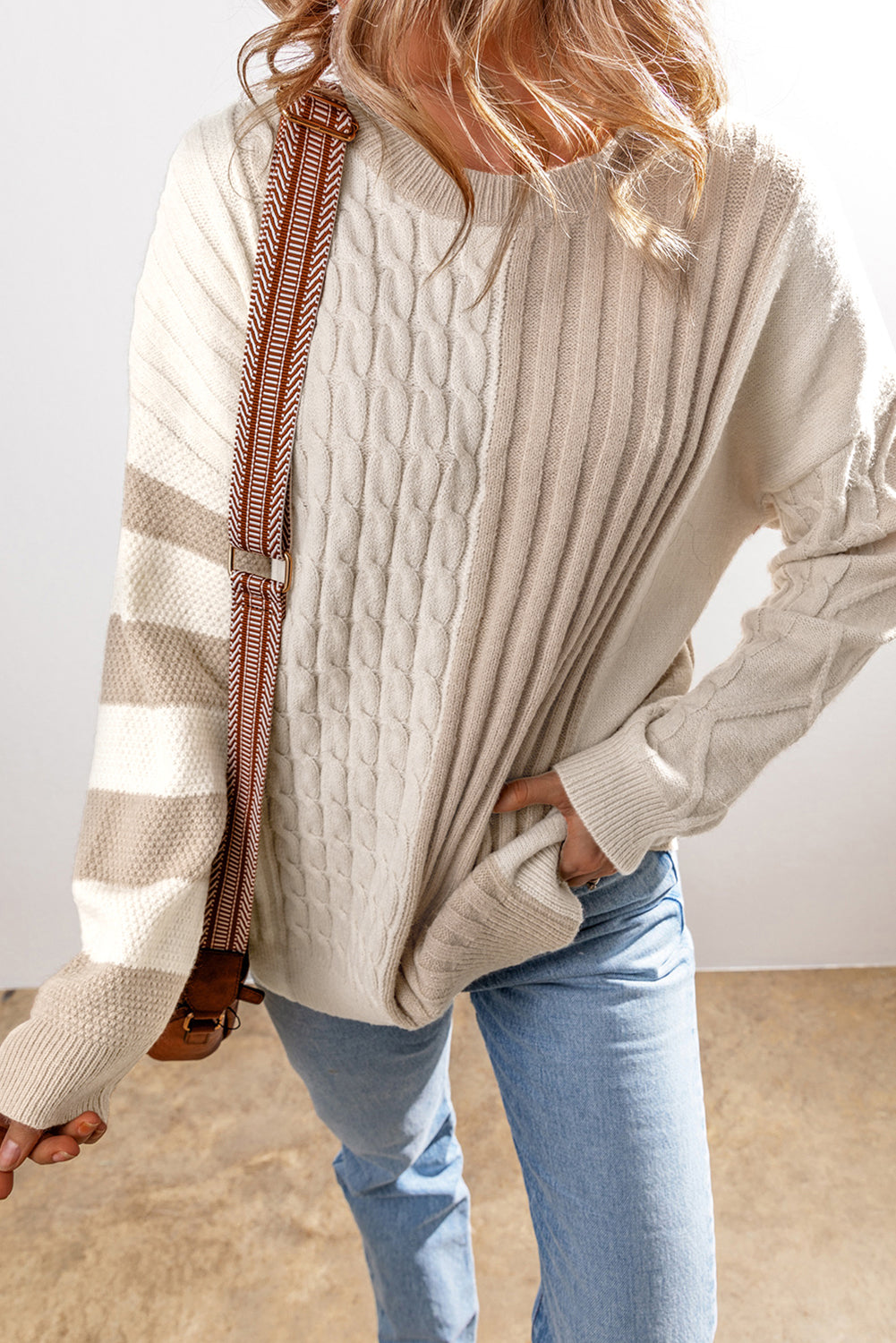 The individual is wearing a Shewin Khaki Cable Knit Colorblock Crew Neck Drop Shoulder Sweater, complemented by a shoulder bag and light blue jeans.