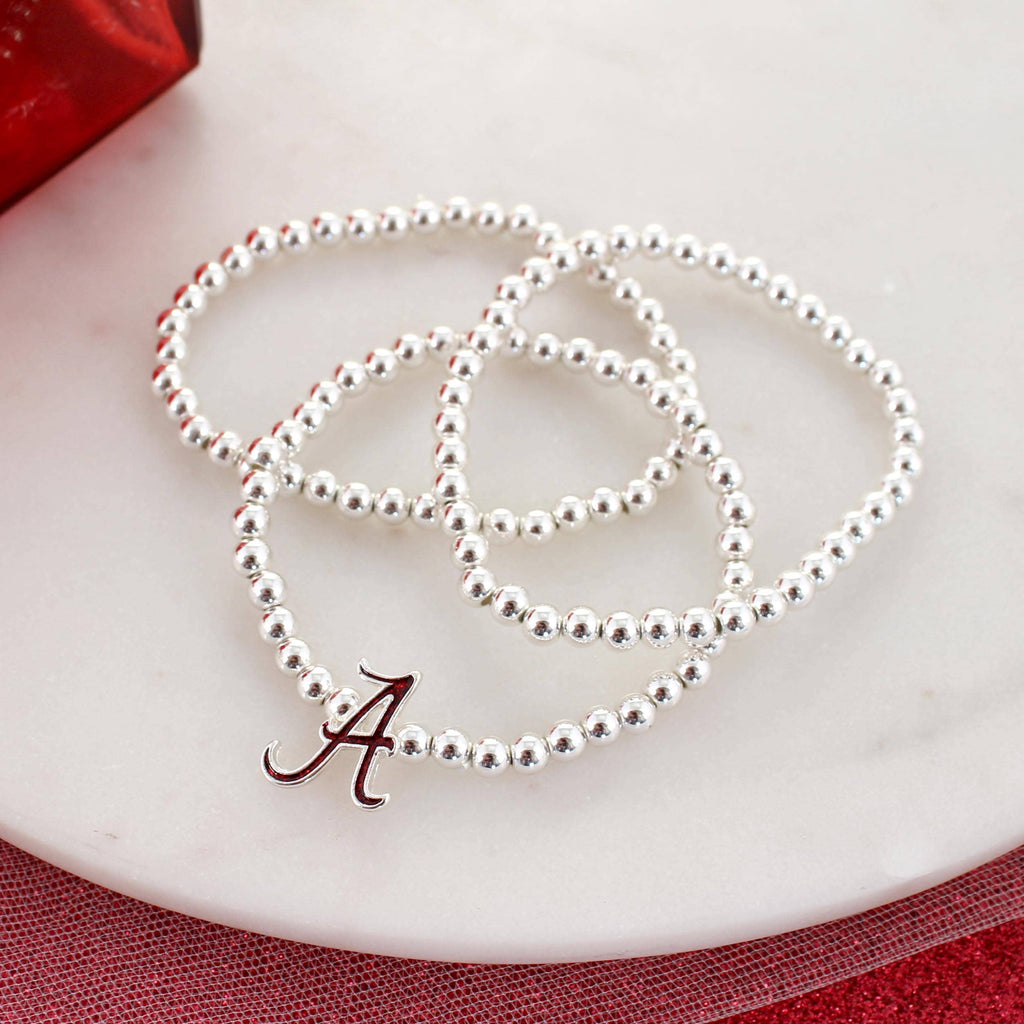 Three silver beaded stretch bracelets from Seasons Jewelry’s Alabama Glitter Enamel Logo Stretch Bracelet Set, each featuring a crimson glitter red “A” charm, are displayed on a white marble surface—making the perfect accessory for any Alabama fan.