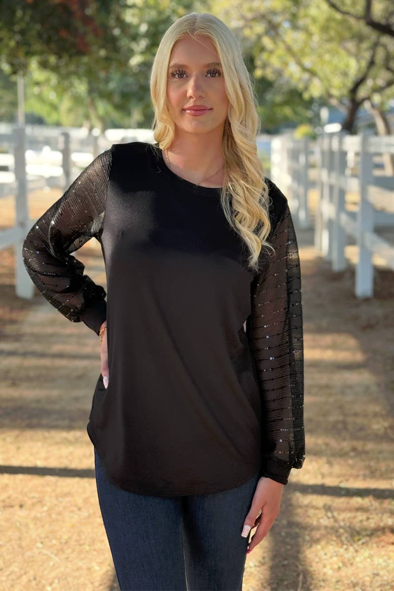 A woman stands outside by a white fence, wearing the 24-Hrs Fashion Sequin Sleeve Blouse—black with sheer balloon sleeves—and dark pants. Sunlight filters through nearby trees in the background.