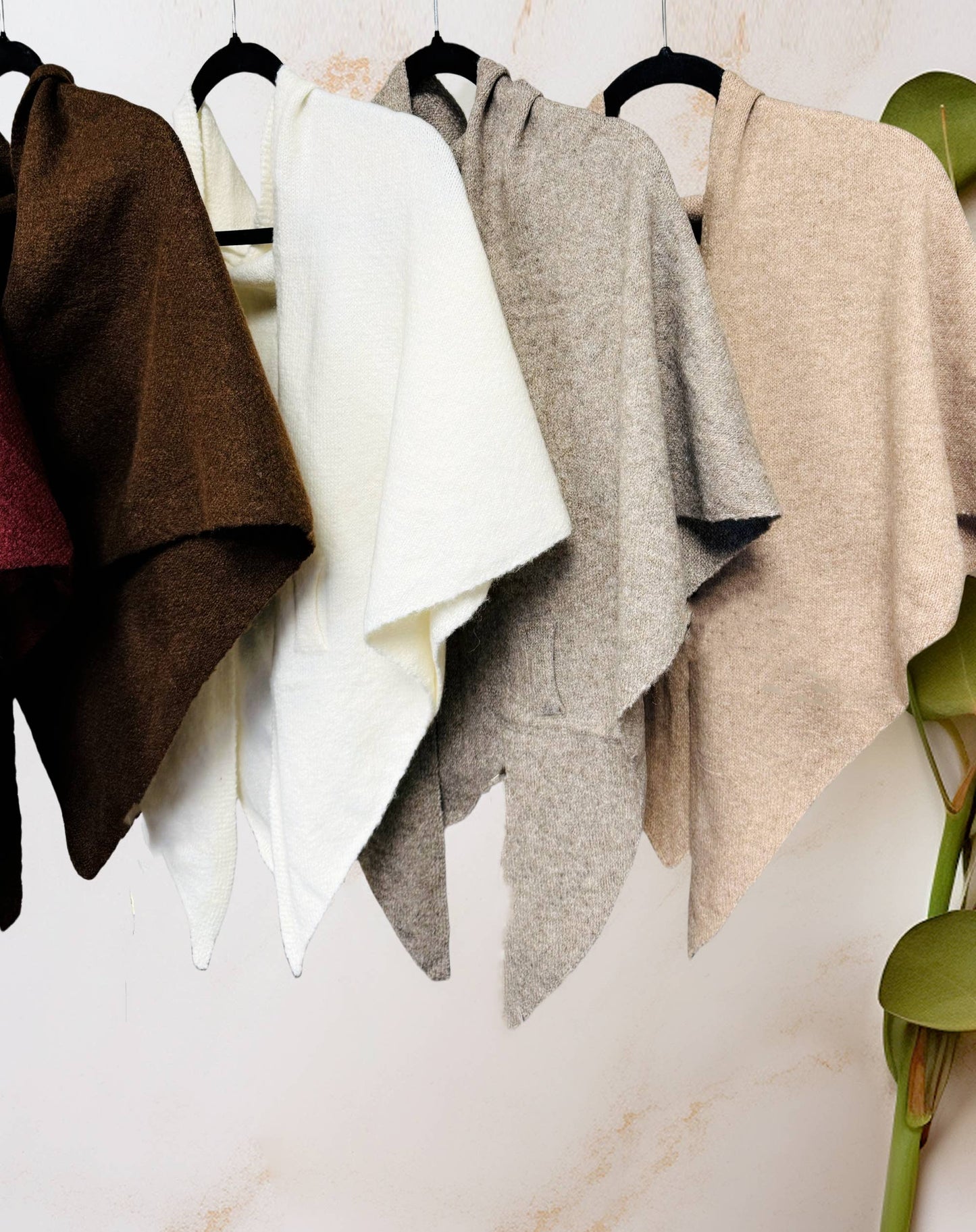 Four JC Sunny Fashion Cashmere-Blend Knit Triangle Shawls with Cross-Front Design in brown, white, gray, and beige hang on black hangers. Each stylish piece doubles as a cozy scarf. A plant is partially visible on the right side.