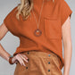 Dressed in Shewin's Patch Pocket Ribbed Knit Short Sleeve Sweater in orange, paired with a button-adorned brown skirt, they exude confidence for transitional weather. Accessories include a long necklace and bracelet, enhancing the look as they stand with a hand on their hip.