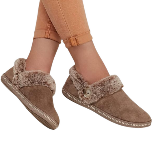 Someone is comfortably wearing the Cozy Campfire Slipper in Taupe by Skechers, lined with faux fur, and paired with vibrant orange pants.