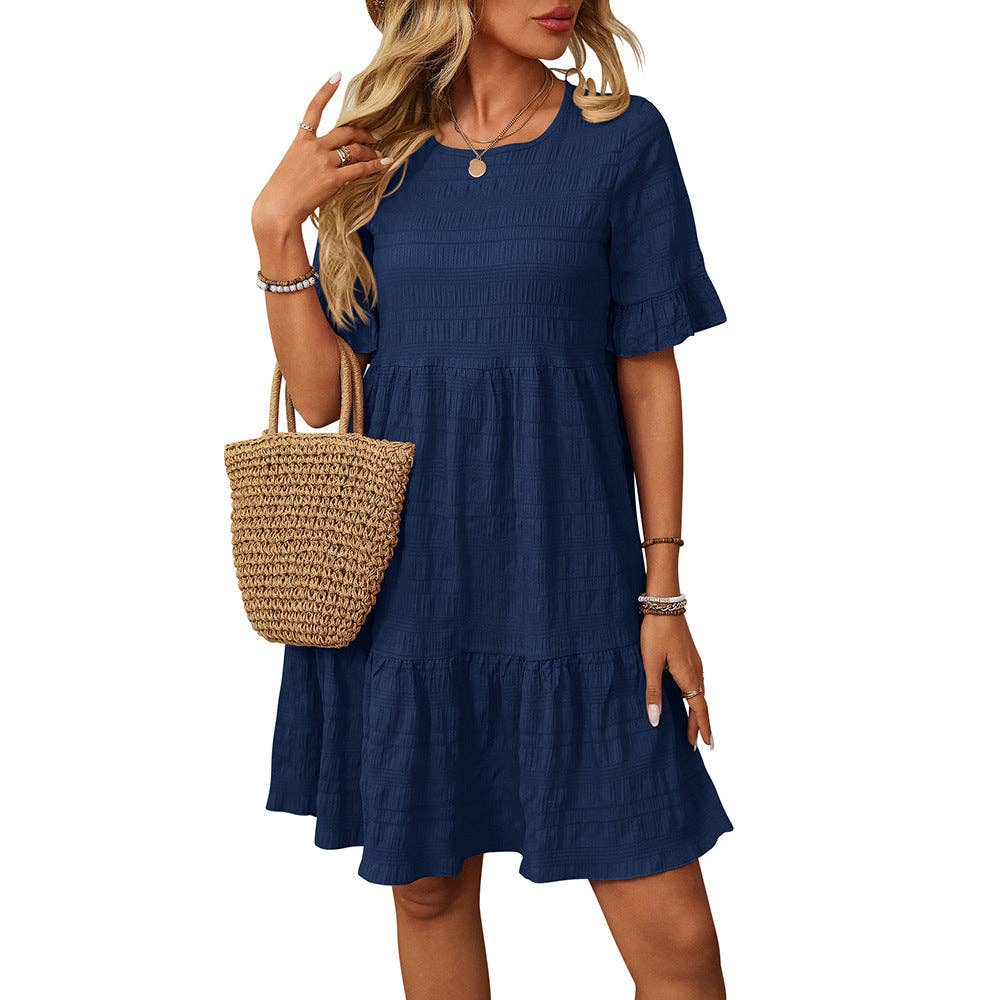 A woman wears the Suhkasana Loose Casual Short-Sleeve Dress in navy, featuring tiered layers. She completes her chic summer look with a woven straw handbag and jewelry—ideal for stylish, warm-weather outings.