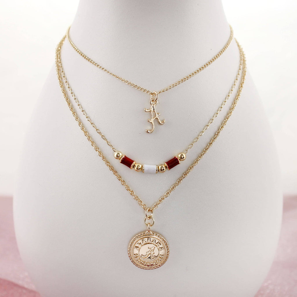 The Alabama Gold & Tube Bead Trio Necklace Set by Seasons Jewelry features three elegant gold-layered chains: a script letter charm, a beaded strand with red and gold beads, and a round pendant engraved with "Alabama Crimson Tide." This collegiate necklace set is the perfect piece of Bama jewelry, beautifully showcased on a white display bust.
