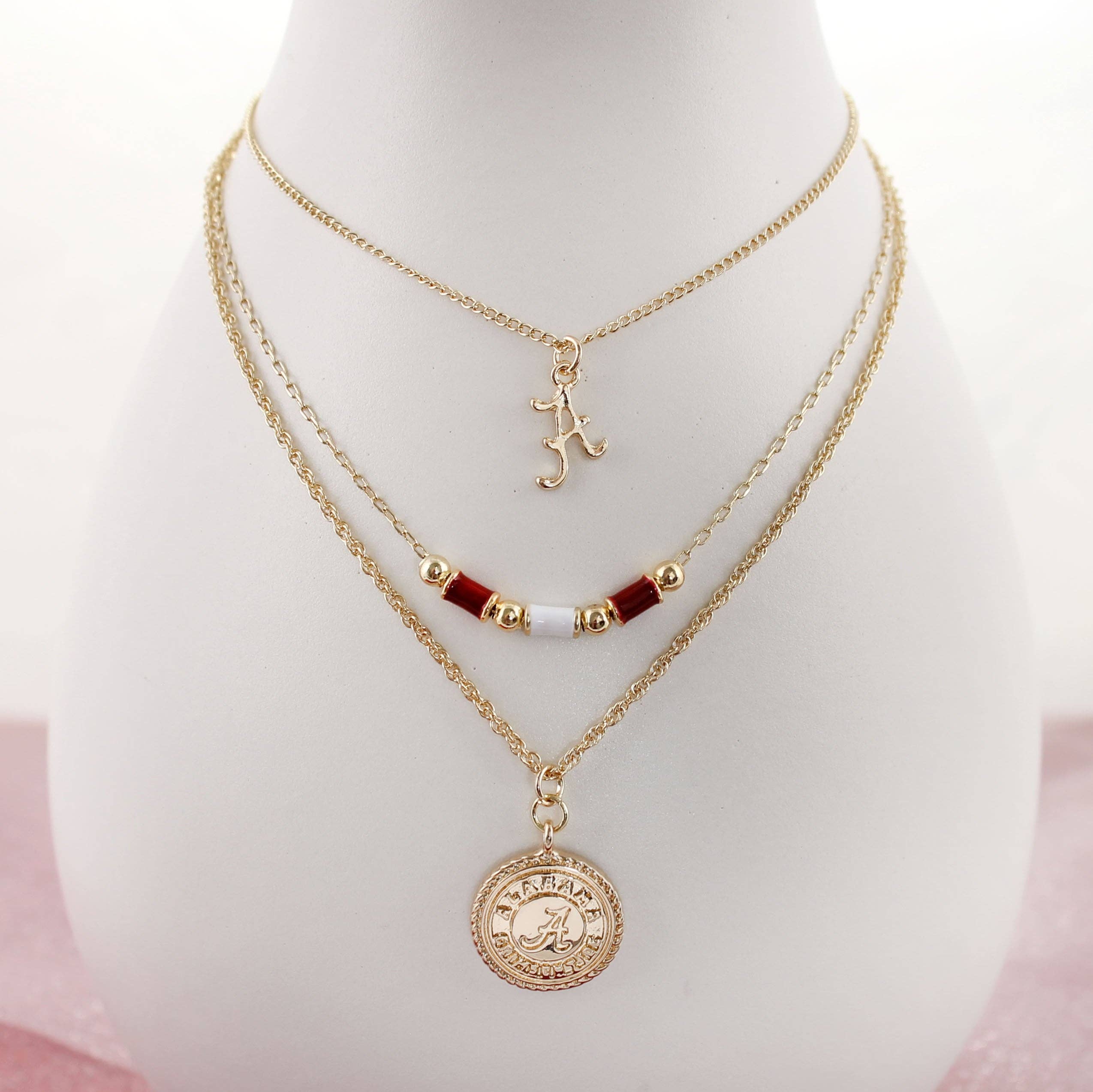 The Alabama Gold & Tube Bead Trio Necklace Set by Seasons Jewelry features three elegant gold-layered chains: a script letter charm, a beaded strand with red and gold beads, and a round pendant engraved with "Alabama Crimson Tide." This collegiate necklace set is the perfect piece of Bama jewelry, beautifully showcased on a white display bust.