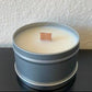 A 4oz Handpoured Soy Wax Candle with a wood wick by Stephanie Henry Candle Co. flickers softly in a gray metal tin of Mahogany Teakwood against a light textured wall.