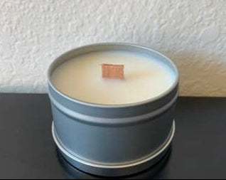 A 4oz Handpoured Soy Wax Candle with a wood wick by Stephanie Henry Candle Co. flickers softly in a gray metal tin of Mahogany Teakwood against a light textured wall.