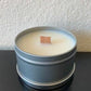 The 4oz Handpoured Soy Wax Candle in a Tin from the Stephanie Henry Candle Company, with a wood wick, sits on a dark surface against a textured white wall.