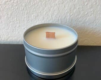 The 4oz Handpoured Soy Wax Candle in a Tin from the Stephanie Henry Candle Company, with a wood wick, sits on a dark surface against a textured white wall.