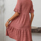 A woman stands indoors with her back to the camera, wearing the Suhkasana Loose Casual Short-Sleeve Dress in rose, featuring textured fabric and a ruffled hem.