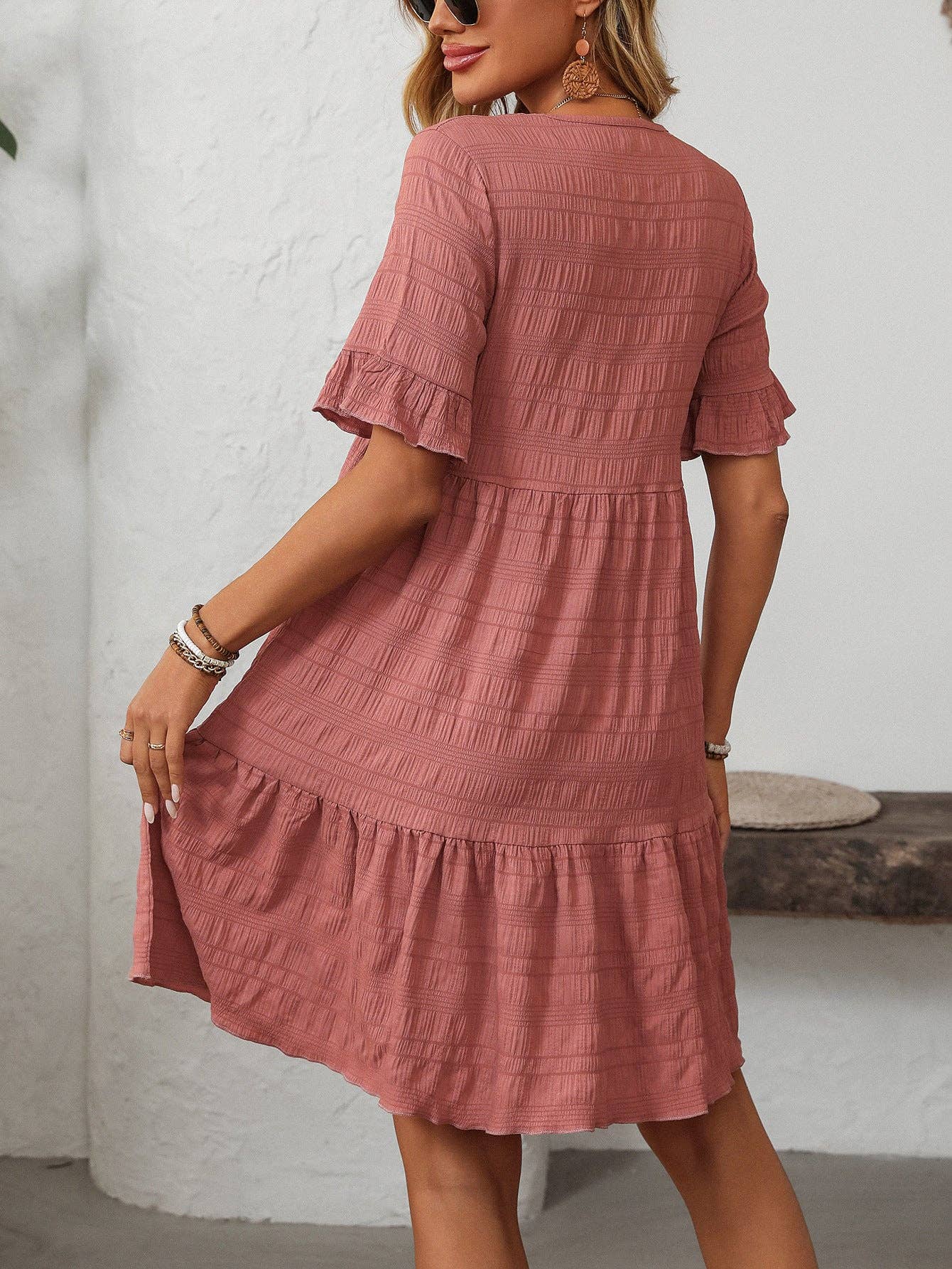 A woman stands indoors with her back to the camera, wearing the Suhkasana Loose Casual Short-Sleeve Dress in rose, featuring textured fabric and a ruffled hem.