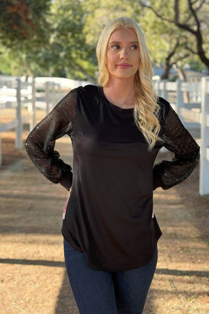 A woman stands outdoors near a white fence, wearing the 24-Hrs Fashion Sequin Sleeve Blouse with sheer balloon sleeves and dark jeans. Sunlight filters through trees in the background.