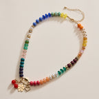 The Lilla Haven Textured Glass Bead Necklace with Charm Trio features vibrant beaded accents on a gold chain, adorned with gold-toned spacers, a gold flower, a heart charm, and a red bead pendant, styled against a neutral background.