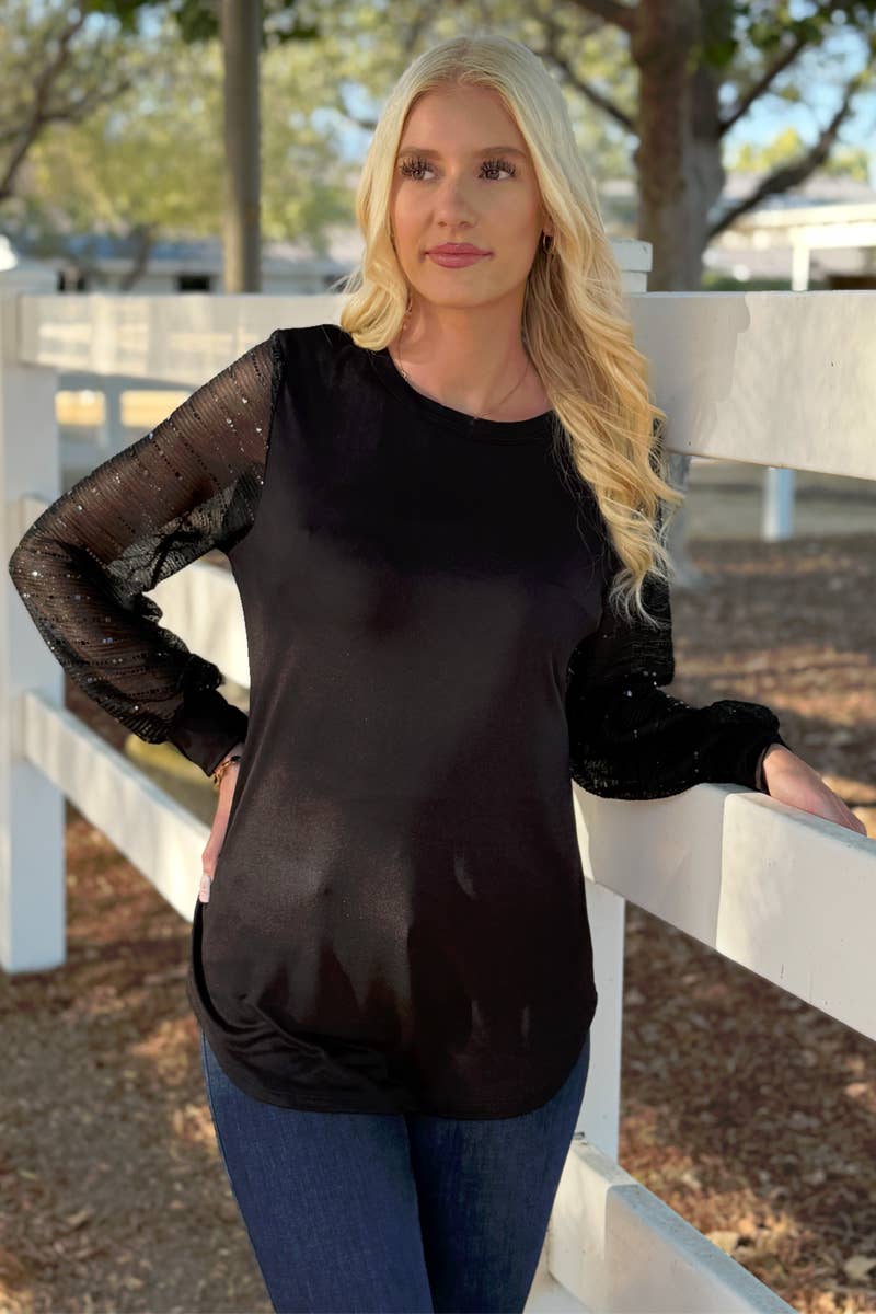 A woman with long blonde hair stands outdoors by a white fence, wearing the 24-Hrs Fashion Sequin Sleeve Blouse and blue jeans. Trees and sunlight can be seen in the background.
