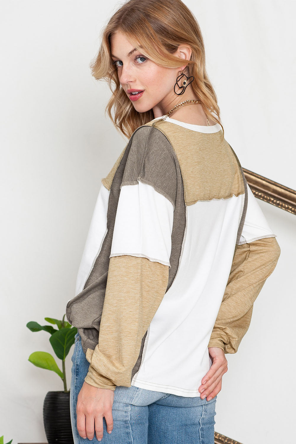 A person with blonde hair is standing and looking over their shoulder. They are wearing a Shewin Khaki Color Block Exposed Seam Long Sleeve Top in shades of white, beige, and gray with a subtle patchwork design, paired with blue jeans.