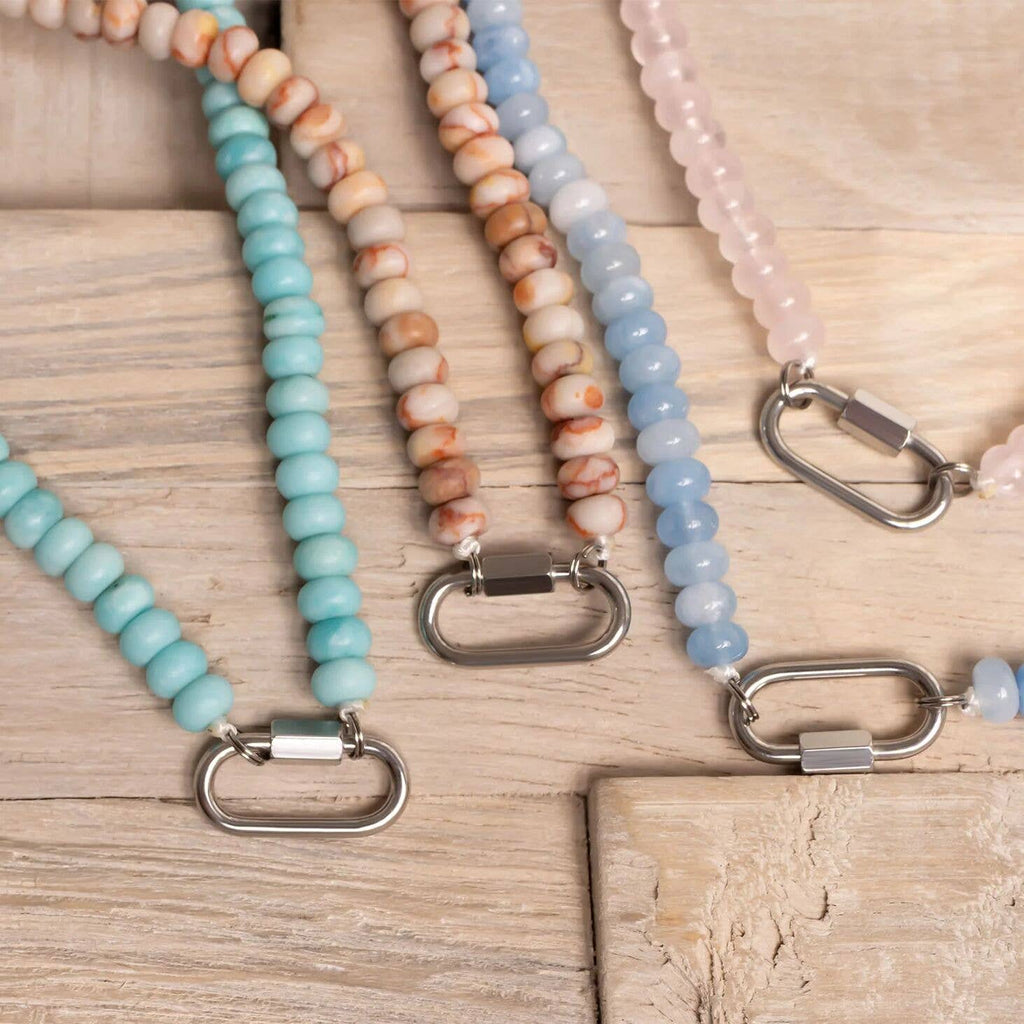 Four WJW necklaces, including the Stainless Steel Chunky Glass Bead Carabiner Necklace in pastel colors, are arranged on a light wooden surface. The collection features both chunky glass beaded designs and sleek stainless steel styles with metal clasps.
