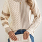 A woman wears a Shewin Khaki Cable Knit Colorblock Crew Neck Drop Shoulder Sweater, holding a vertical line-embossed cream clutch, paired with blue jeans, a gold necklace, and rings.