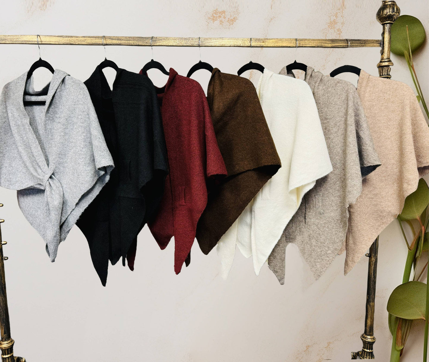 Seven ponchos in gray, black, burgundy, brown, white, light gray, and beige hang on black hangers from a brass rack—ideal for layering with the JC Sunny Fashion Cashmere-Blend Knit Triangle Shawl with Cross-Front Design.