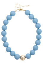 Introducing the Marjorie Game Day Beaded Necklace by Canvas Style—a bold statement piece featuring round blue beads and a single oversized gold bead at the center, finished with a gold clasp and an adjustable chain for versatile wear on any occasion.
