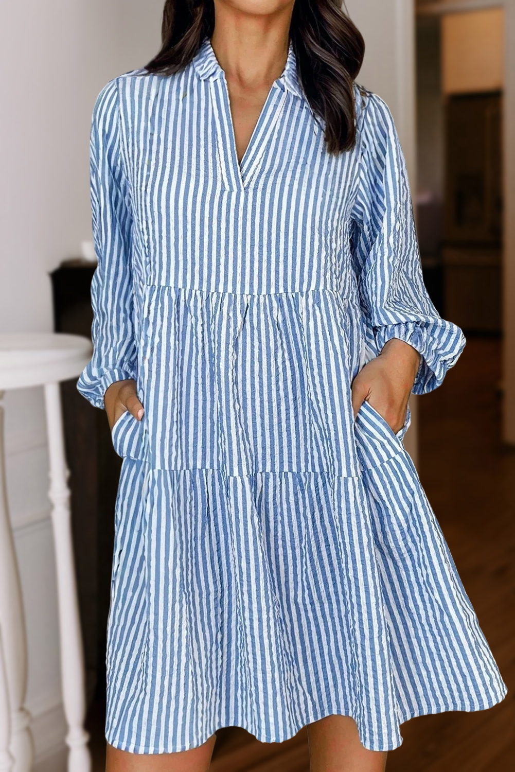 A woman stands indoors wearing the Aapparella Stripe Tiered Pocket 3/4 Sleeve Shift Dress, featuring blue and white vertical stripes, a loose fit, and convenient pockets.