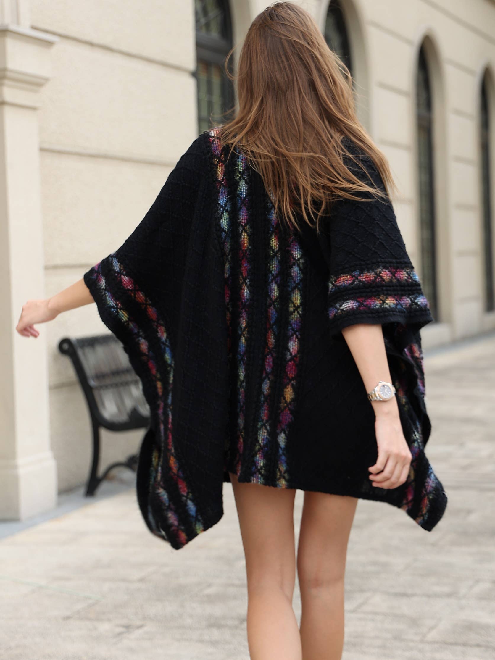 A woman with long brown hair wears the JC Sunny Fashion Black Rainbow Trim Pocket Ruana while walking outdoors near a beige building and a black bench.