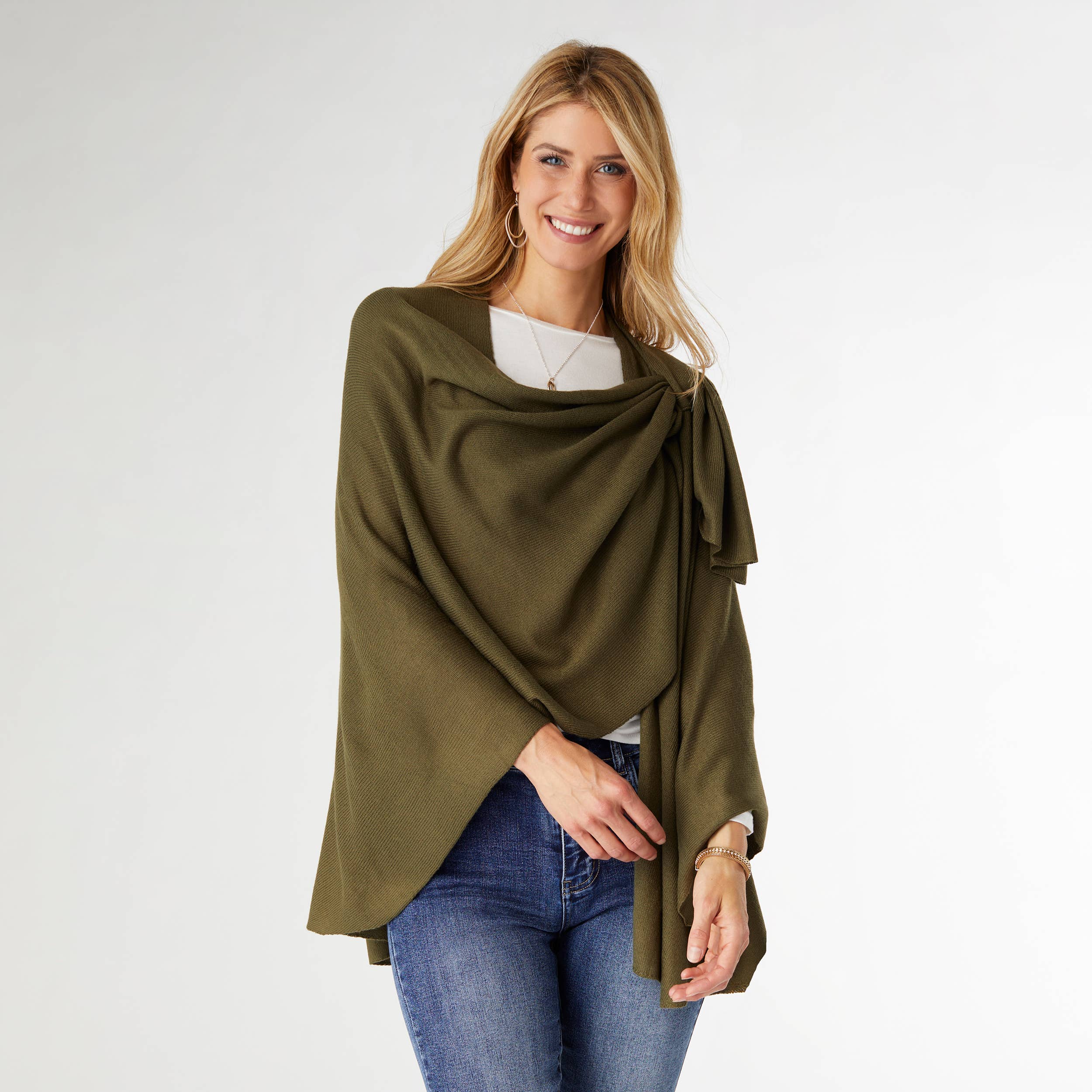 A woman models the COCO + CARMEN Kiara Pull Thru Wrap in green, styled over a white top and blue jeans, smiling against a plain light background.