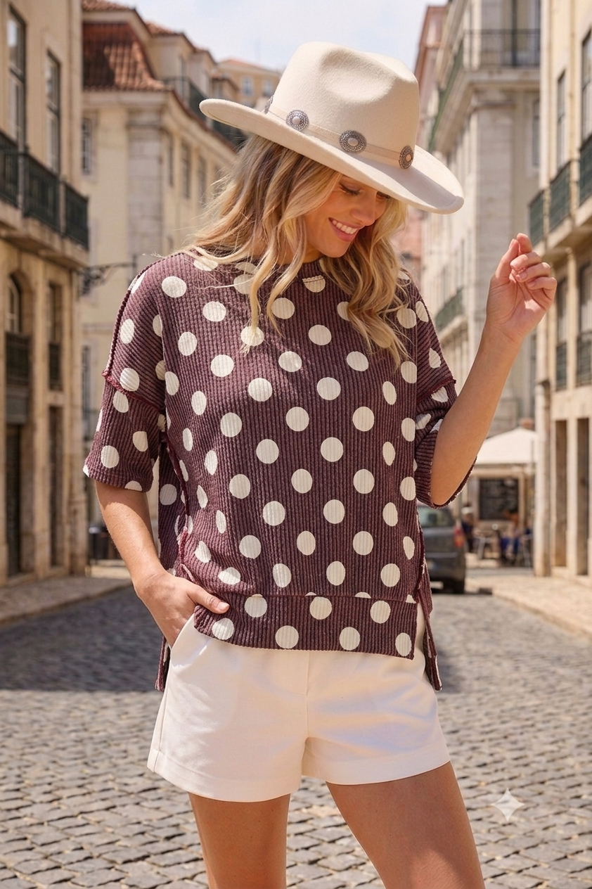 A woman wearing the Celeste Clothing Women’s Polka Dot Rib Knit Top in a relaxed fit, paired with a cream hat and white shorts, stands on a cobblestone street between old buildings on a sunny day.