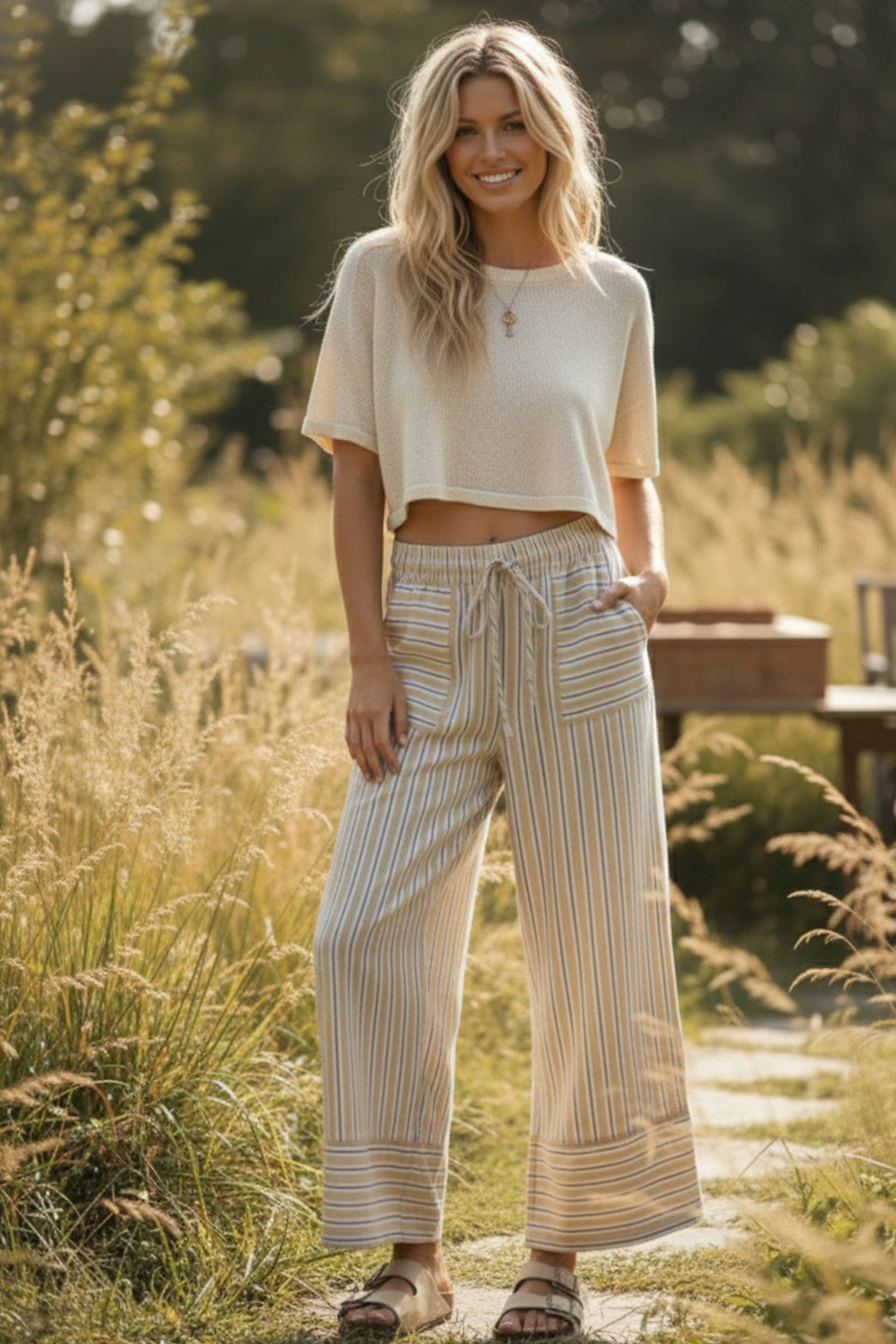 A woman stands outdoors on a path in Sewn and Seen's Woven Striped Wide Leg Pants paired with a short-sleeve beige top and sandals, surrounded by tall grass and trees for a coastal-inspired backdrop.