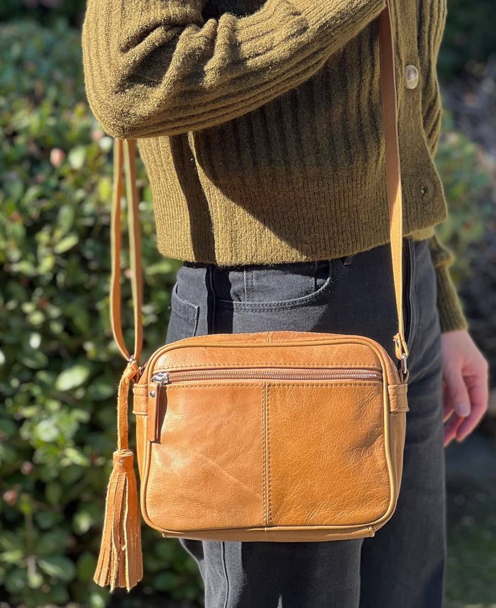 Outdoors, someone in a green sweater and black pants carries the Roma Leathers, Inc. Cowhide Leather Crossbody, featuring an adjustable shoulder strap and tassel.