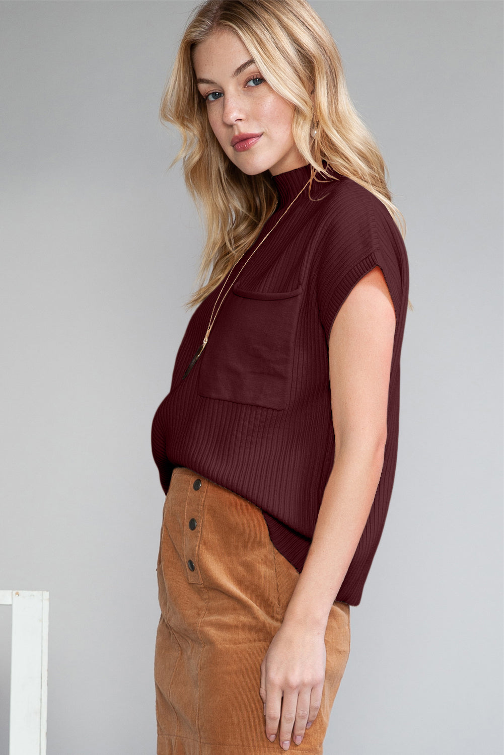 A blonde woman in a Shewin Patch Pocket Ribbed Knit Short Sleeve Sweater and a tan skirt stands confidently against a plain backdrop, embodying the essence of transitional weather.