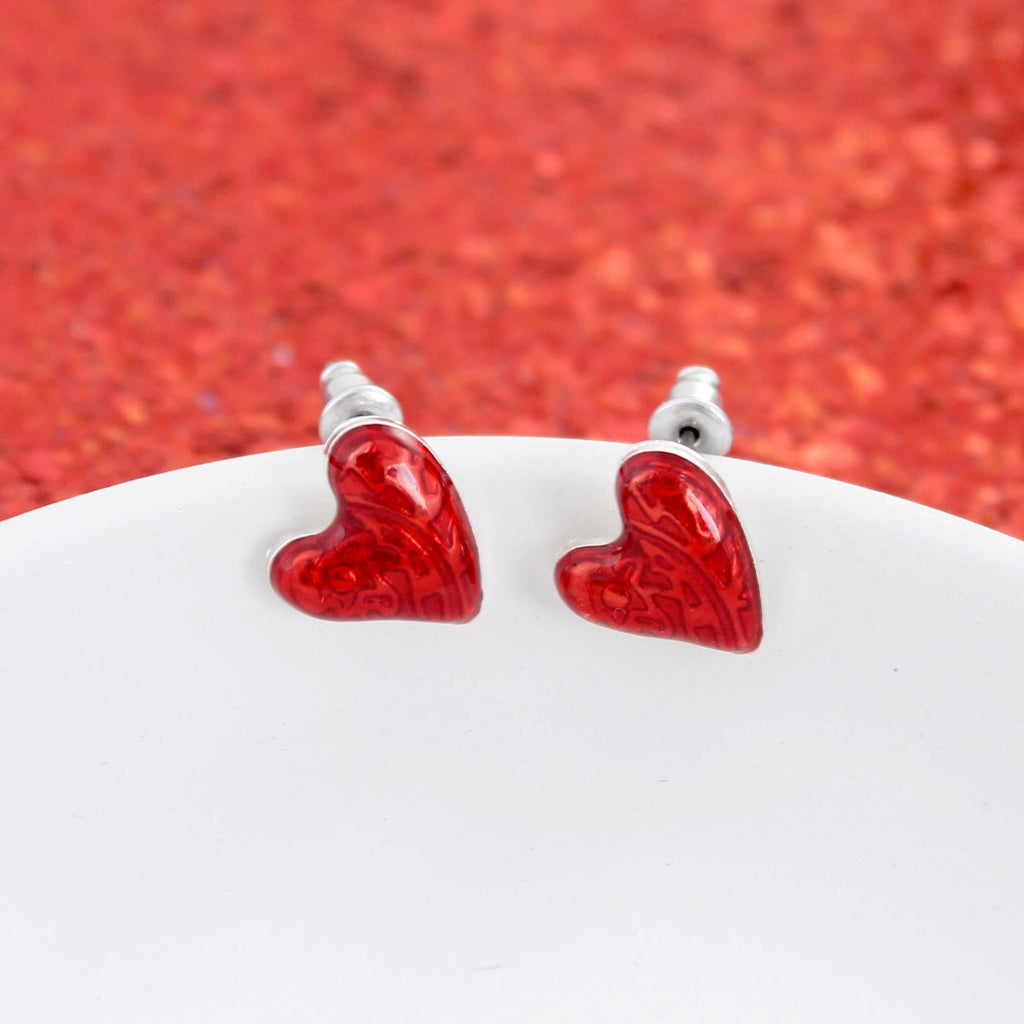 A pair of Puffy Heart Valentine’s Day Stud Earrings from Seasons Jewelry, featuring small red enamel hearts, displayed on a white surface with a red textured background—perfect as Valentine’s Day jewelry.