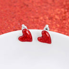 A pair of Puffy Heart Valentine’s Day Stud Earrings from Seasons Jewelry, featuring small red enamel hearts, displayed on a white surface with a red textured background—perfect as Valentine’s Day jewelry.