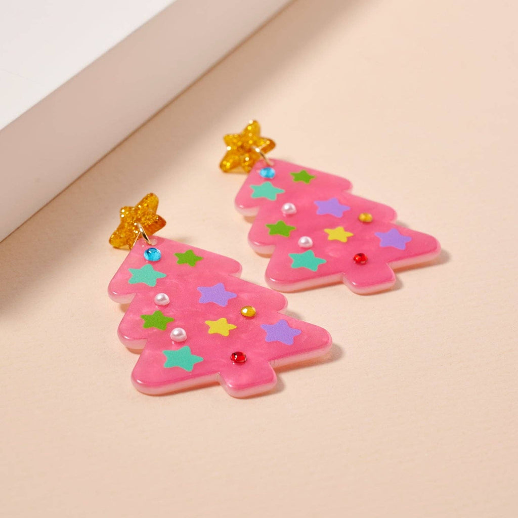 Add some specifics to highlight the product name and brand. Here’s a rewritten version: The Glittery Christmas Tree Drop Earrings by Lilla Haven are crafted from sparkling pink acrylic and adorned with colorful star and bead accents, making them the perfect holiday statement piece. Each earring is topped with a cheerful yellow star, beautifully displayed on a light beige surface.