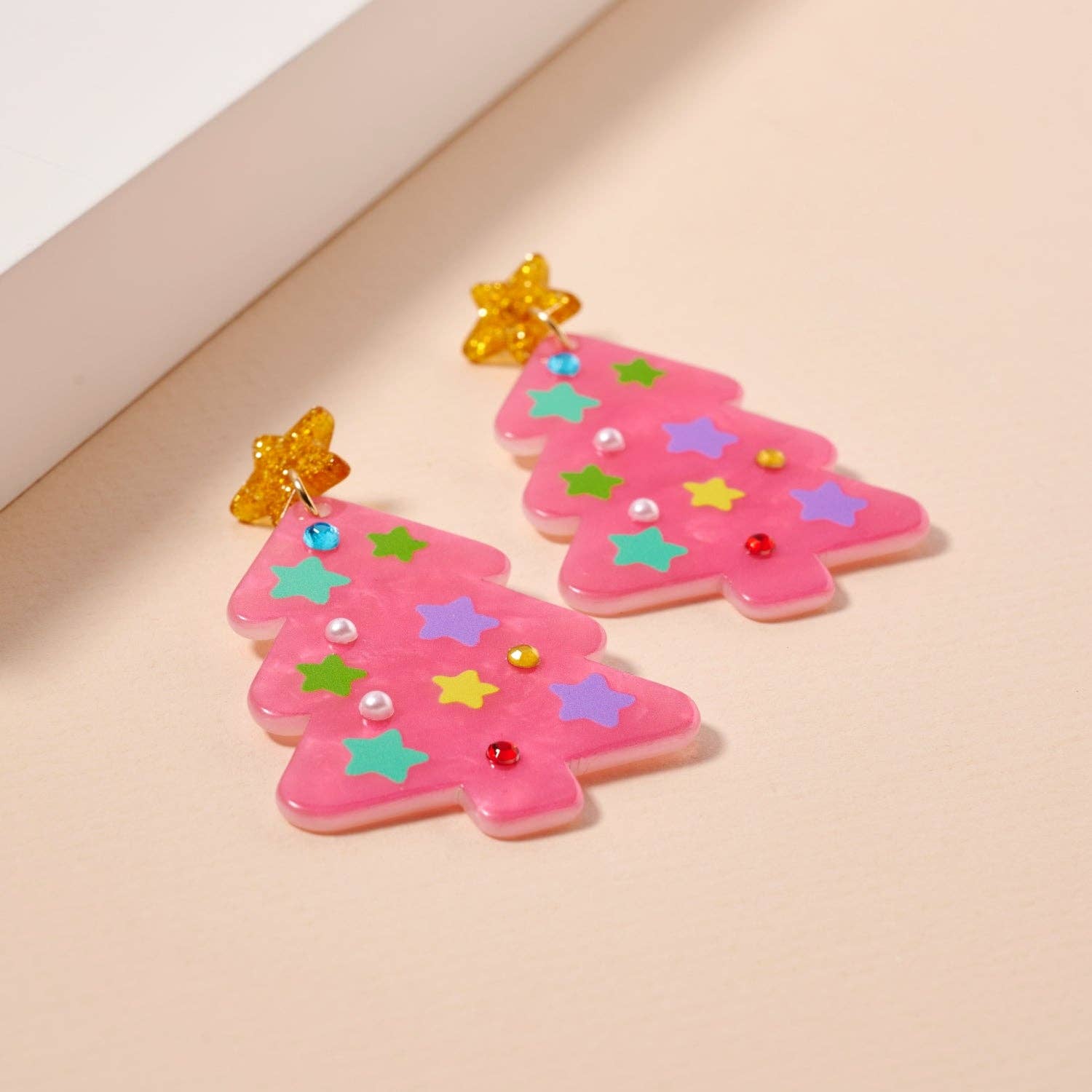 Add some specifics to highlight the product name and brand. Here’s a rewritten version: The Glittery Christmas Tree Drop Earrings by Lilla Haven are crafted from sparkling pink acrylic and adorned with colorful star and bead accents, making them the perfect holiday statement piece. Each earring is topped with a cheerful yellow star, beautifully displayed on a light beige surface.
