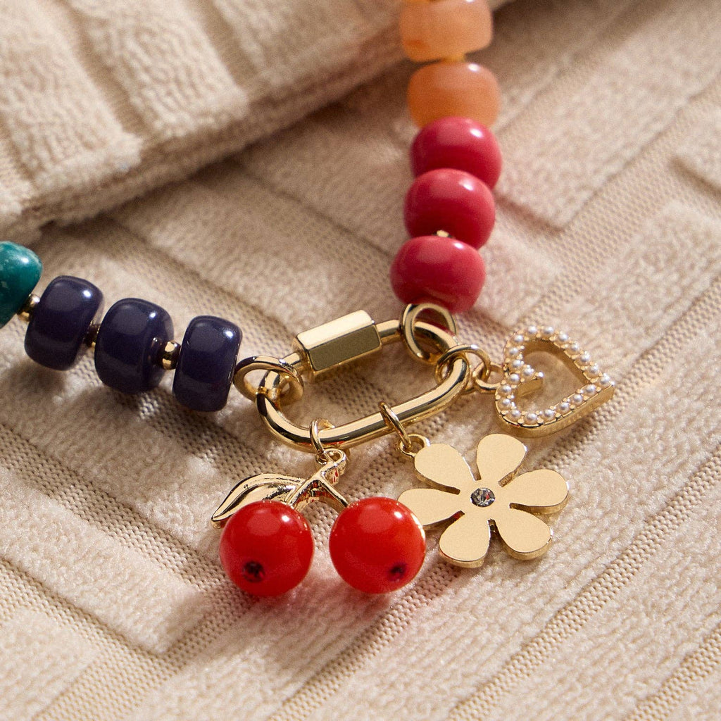 Close-up of the Lilla Haven Textured Glass Bead Necklace with Charm Trio, featuring multicolored glass beads and gold-tone spacers, adorned with a heart, a flower, and two red cherry charms, displayed on textured beige fabric.