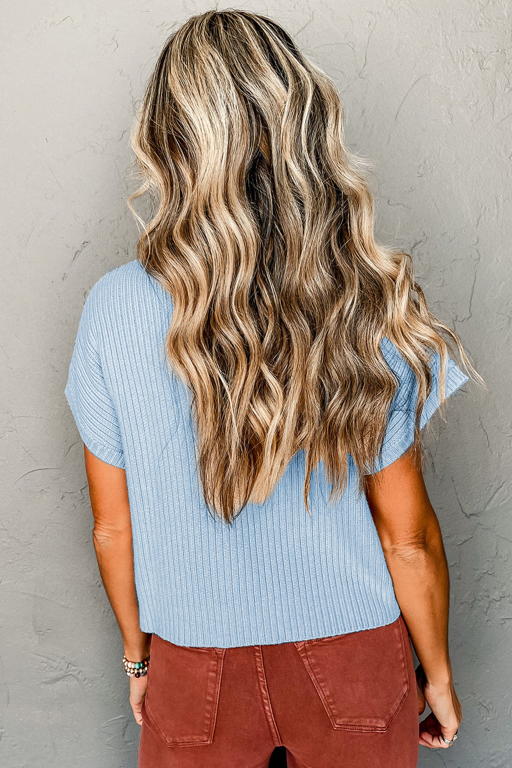 Person with long wavy blonde hair in a Shewin Patch Pocket Ribbed Knit Short Sleeve Sweater and brown pants, perfectly dressed for transitional weather, facing a textured gray wall.