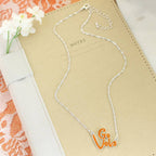 The Tennessee Slogan Necklace by Seasons Jewelry, featuring a silver chain and an orange glitter enamel pendant, is displayed on a tan notebook labeled "NOTES," beside white flowers and orange lace—making it the perfect game day accessory for any fan.