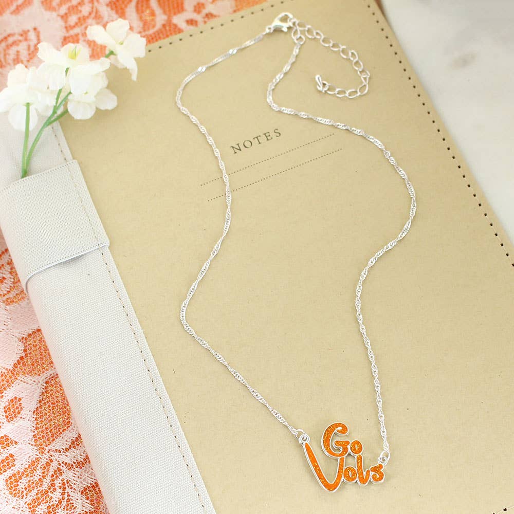 The Tennessee Slogan Necklace by Seasons Jewelry, featuring a silver chain and an orange glitter enamel pendant, is displayed on a tan notebook labeled "NOTES," beside white flowers and orange lace—making it the perfect game day accessory for any fan.