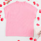 The Shewin Patch Pocket Ribbed Knit Short Sleeve Sweater in pink lies flat amidst scattered rose petals on a white background, offering charm and elegance for transitional weather with its soft knit fabric.