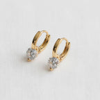 A pair of Ellemadera Modern Geometric Zirconia Drop Earrings, each adorned with a single round clear stone and set in a contemporary gold hoop design, displayed on a white textured surface.
