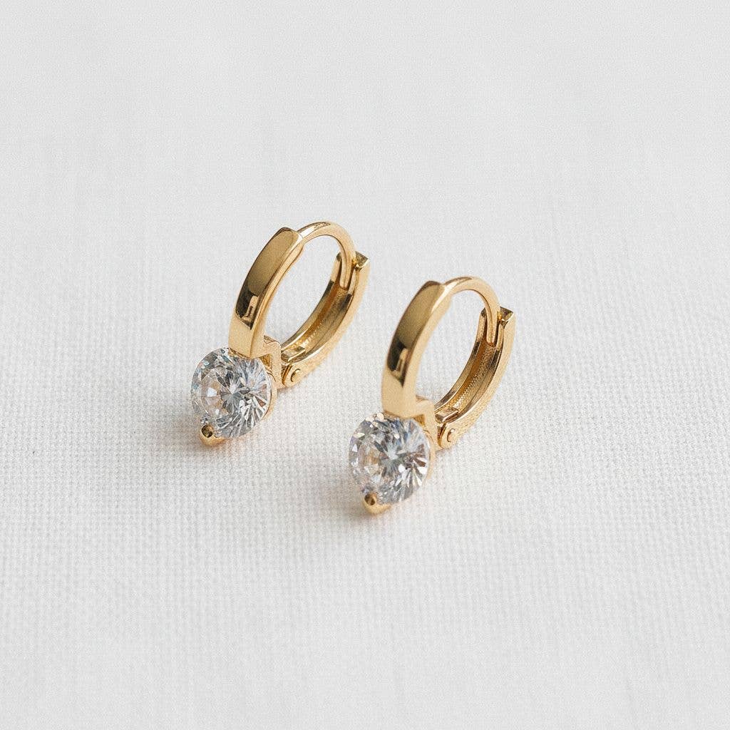 A pair of Ellemadera Modern Geometric Zirconia Drop Earrings, each adorned with a single round clear stone and set in a contemporary gold hoop design, displayed on a white textured surface.