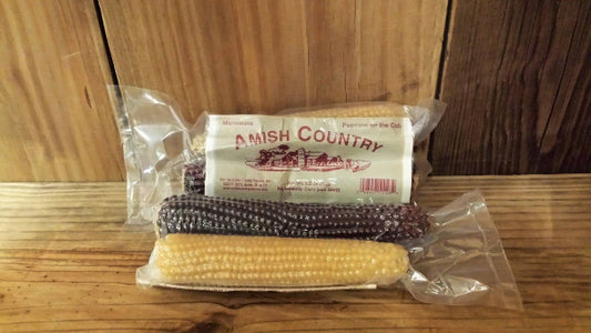 Two vacuum-sealed Twin Microwave Popcorn Cobs from Amish Country Popcorn, with colorful kernels, are displayed on a wooden surface against a wooden background.