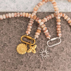 WJW's Stainless Steel Chunky Glass Bead Carabiner Necklace, adorned with sun and bee charms, is showcased alongside another stainless steel necklace featuring a sunburst charm; both are displayed on a pebbled surface background.