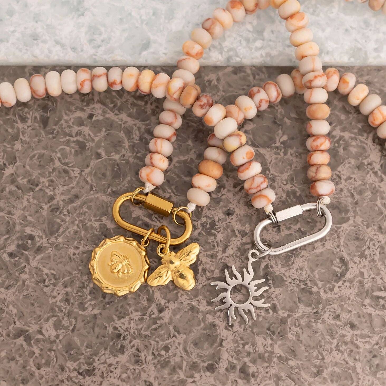 WJW's Stainless Steel Chunky Glass Bead Carabiner Necklace, adorned with sun and bee charms, is showcased alongside another stainless steel necklace featuring a sunburst charm; both are displayed on a pebbled surface background.