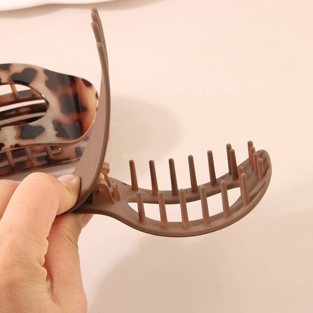 A hand holding an open ACCITY Oval Flat Hair Clip for Women in brown tortoise shell with a second leopard print clip in the background on a beige surface.