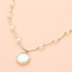 The Round Pearl Pendant Station Necklace by Sensibling Corp. features a delicate beaded chain adorned with small white pearls spaced along its length and a larger, round pearl pendant set in gold. The necklace includes an adjustable extender for the perfect fit and is shown against a light peach background.