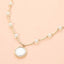 The Round Pearl Pendant Station Necklace by Sensibling Corp. features a delicate beaded chain adorned with small white pearls spaced along its length and a larger, round pearl pendant set in gold. The necklace includes an adjustable extender for the perfect fit and is shown against a light peach background.