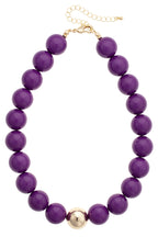 The Marjorie Game Day Beaded Necklace by Canvas Style features an oversized, chunky design with large round purple beads, a central gold accent bead, and is finished with a gold clasp and adjustable chain.